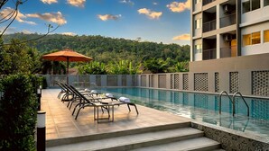 A pool with lounge chairs and an umbrella, surrounded by a fence and greenery, with a building in the background.