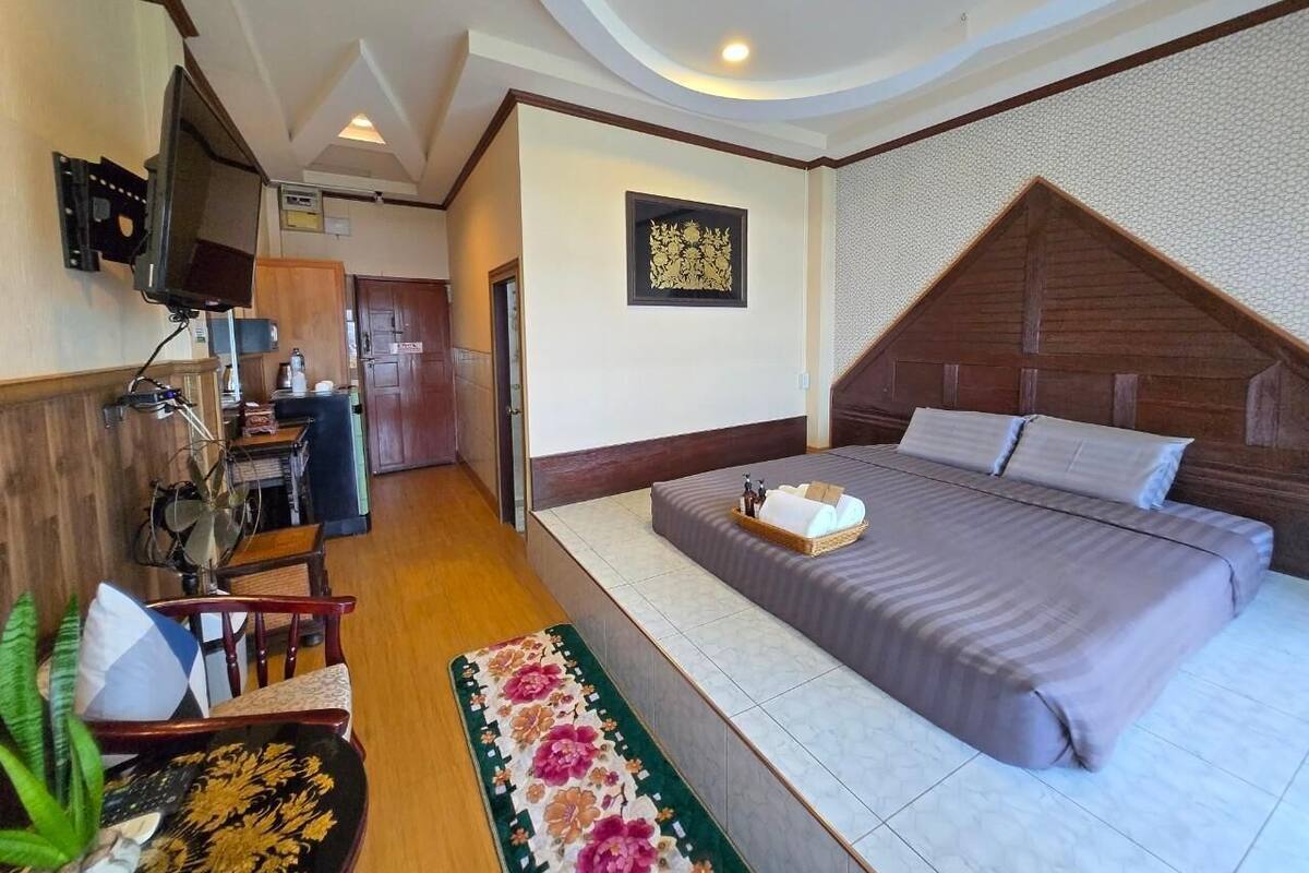 A bedroom with a bed, a TV mounted on the wall, a wooden headboard, and a small kitchenette.