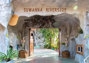 A cave-like entrance with a sign reading 'SUWANNA RIVERSIDE' and statues on wooden pedestals.