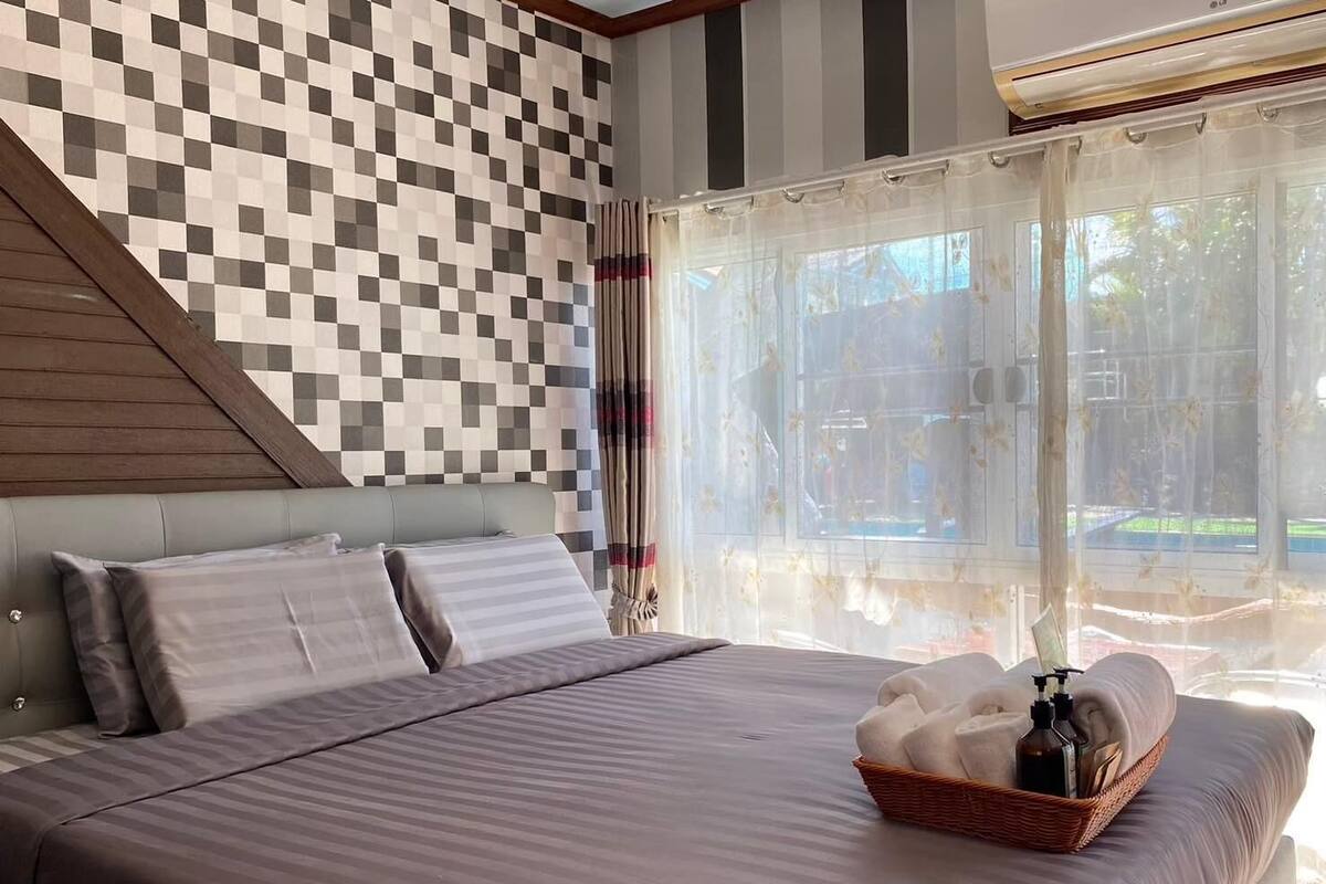 A bedroom with a checkered wall, a bed with pillows, a window with sheer curtains, and a basket with towels and toiletries.