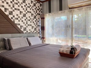 A bedroom with a checkered wall, a bed with pillows, a window with sheer curtains, and a basket with towels and toiletries.