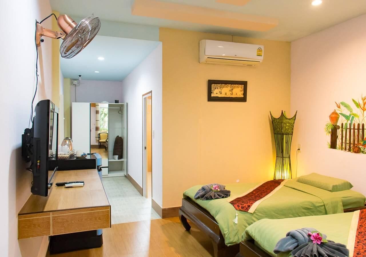 A hotel room with a desk, two beds, a standing fan, an air conditioner, and a hallway leading to a bathroom.