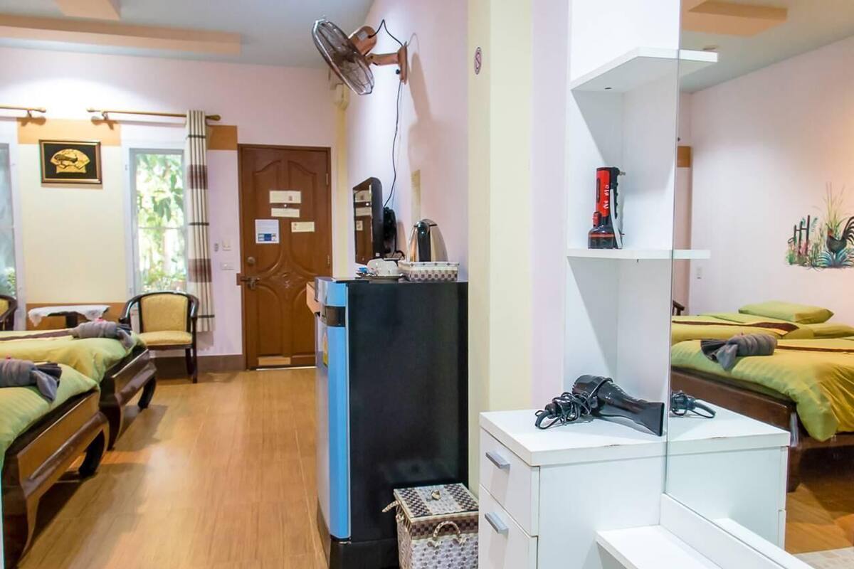 A hotel room with two beds, a desk, a chair, a refrigerator, a fan, and a small cabinet.