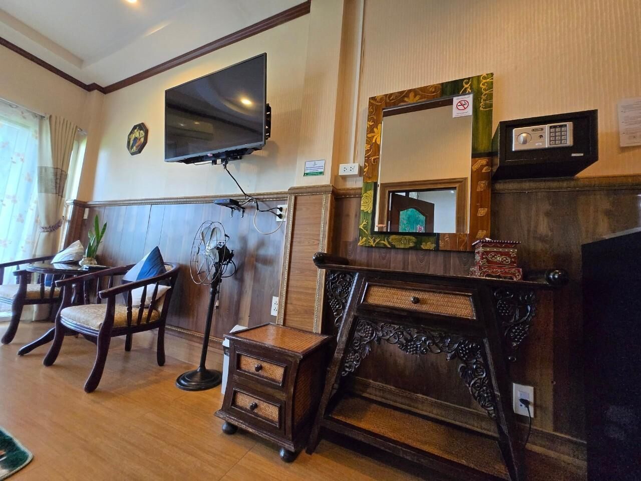 Una habitación con televisión instalada en la pared, una mesa pequeña con sillas, un espejo decorativo y un armario de madera.