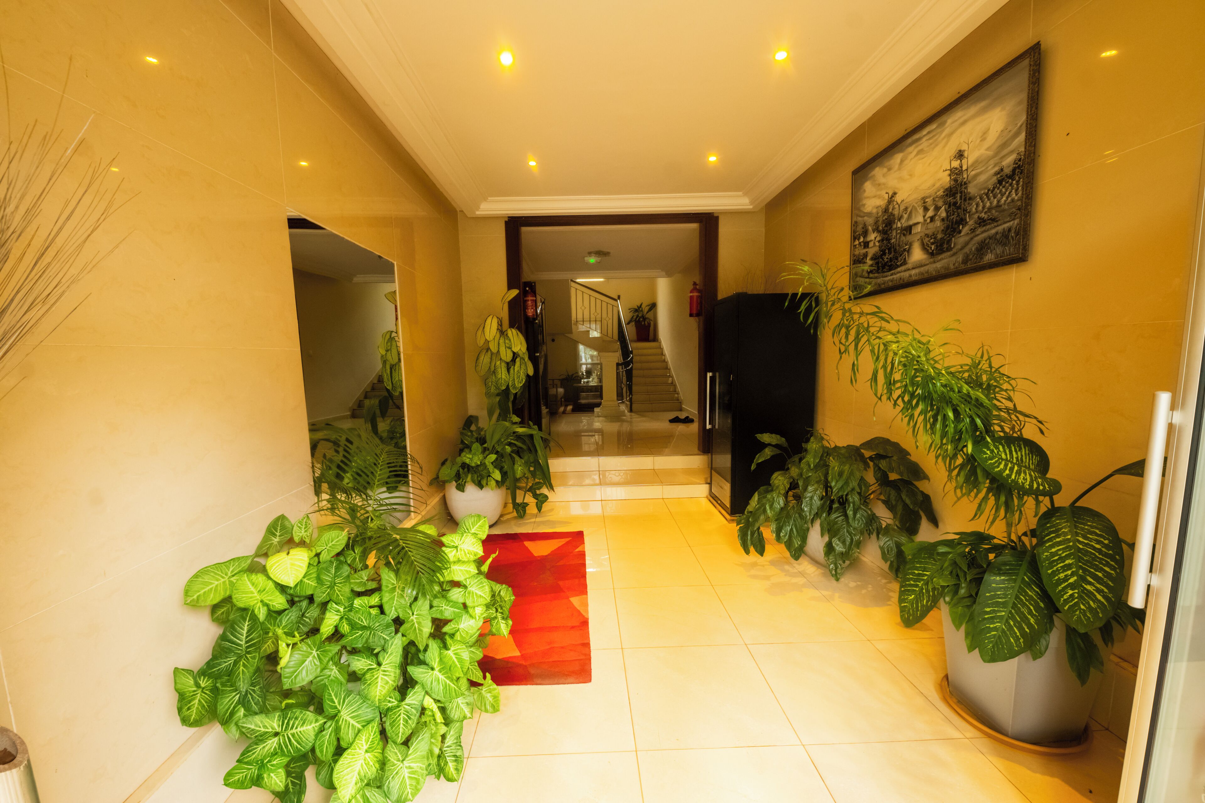 Un hall d’hôtel avec un sol carrelé, des plantes en pot, une moquette rouge et un escalier menant aux étages supérieurs.