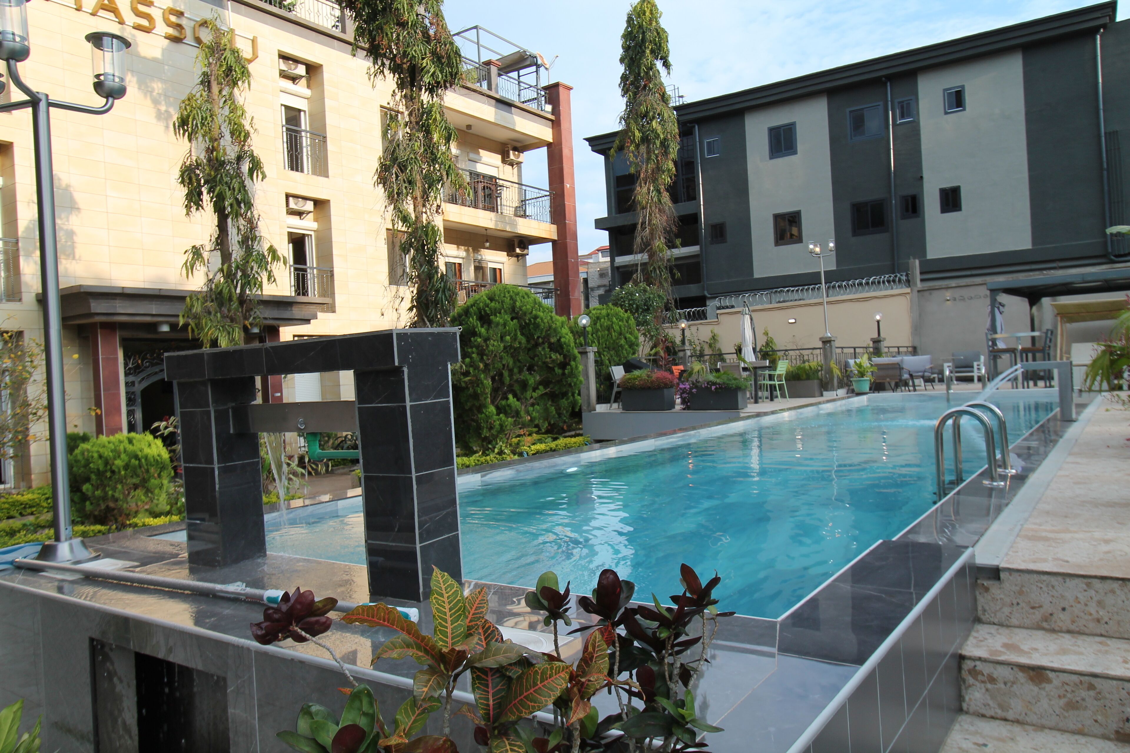 A swimming pool with a tiled deck, surrounded by greenery and modern buildings.