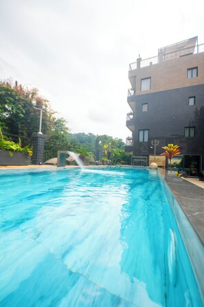 A swimming pool with a water feature, surrounded by a modern building with balconies and a view of trees. - Residence Massou Bastos Golf (Yaoundé)