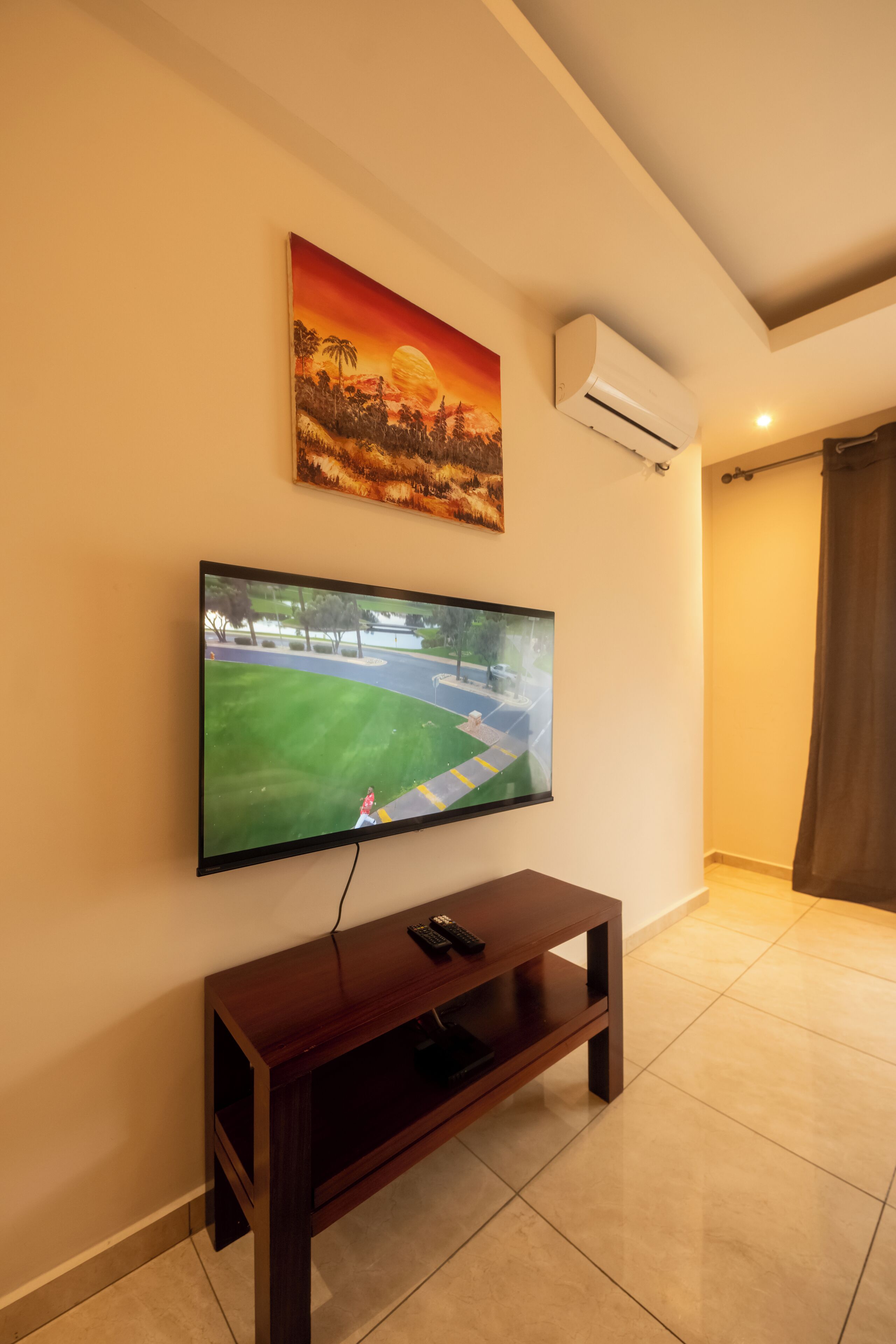 A living room with a flat-screen TV mounted on the wall, a wooden entertainment unit below, and a painting above the TV.