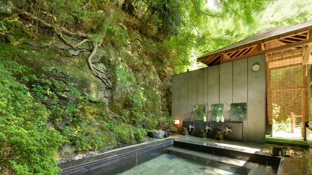 Un onsen japonais traditionnel, avec une source d’eau chaude claire, des marches en pierre menant à l’eau, une structure en bois dot ée de portes coulissantes et un mur de pierre recouvert de mousse.