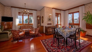 Une salle à manger avec une table dressée pour quatre personnes, entourée de chaises, d’un lustre et d’une grande fenêtre ornée de rideaux.