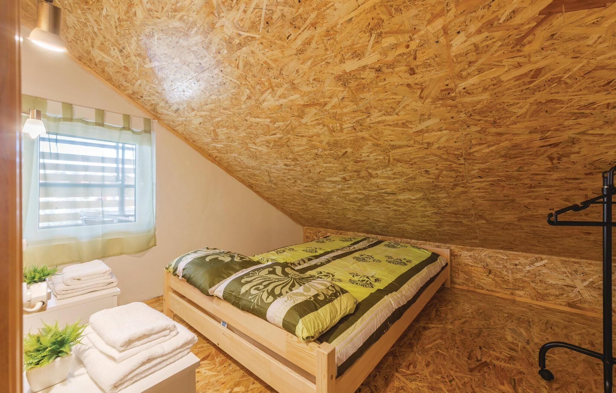 A bedroom with a sloped ceiling, a bed with a patterned bedspread, a window with blinds, and a bedside table with towels and a plant.
