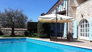Una piscina con muro de piedra, un porche cubierto con sillas y un edificio de piedra con contraventanas azules.