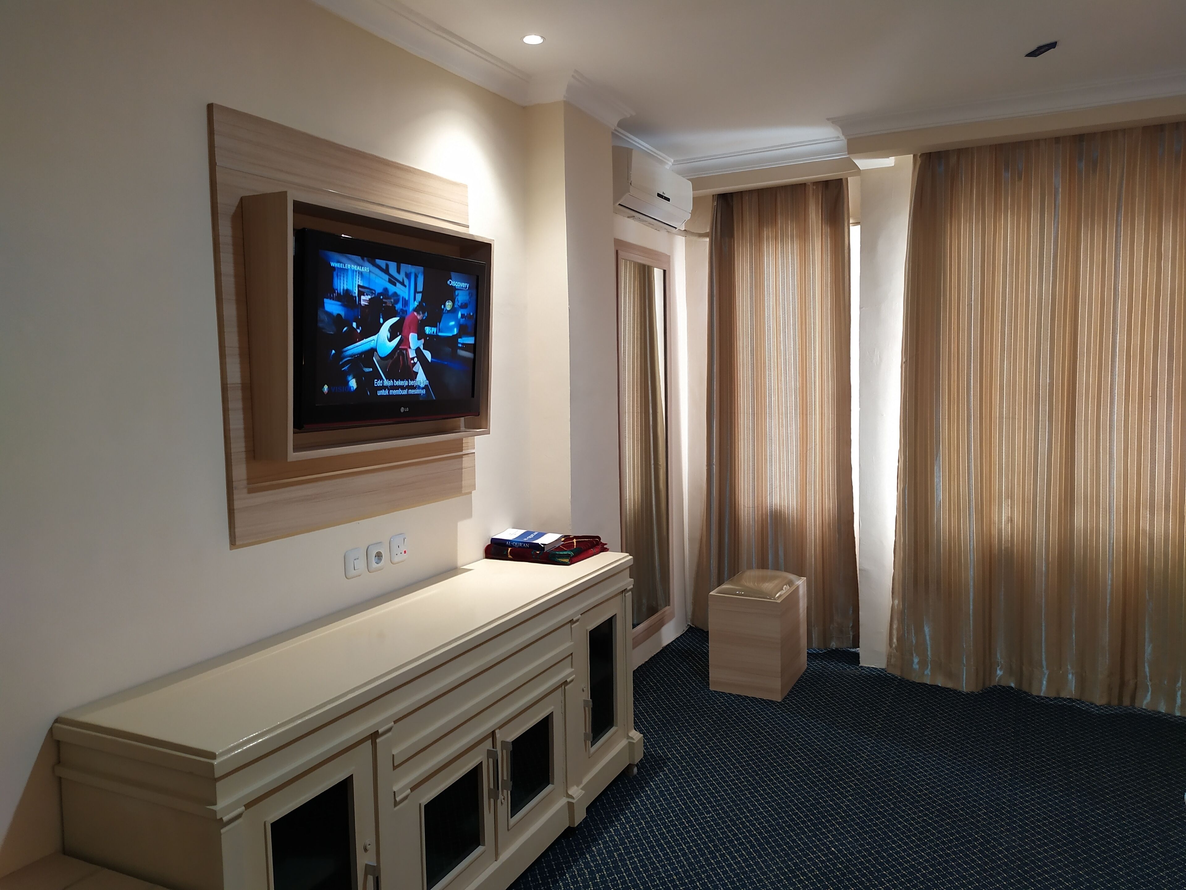 Une chambre d’hôtel équipée d’un téléviseur à écran plat fixé au mur, d’un minibar, d’une petite poubelle et d’une fenêtre avec des rideaux.