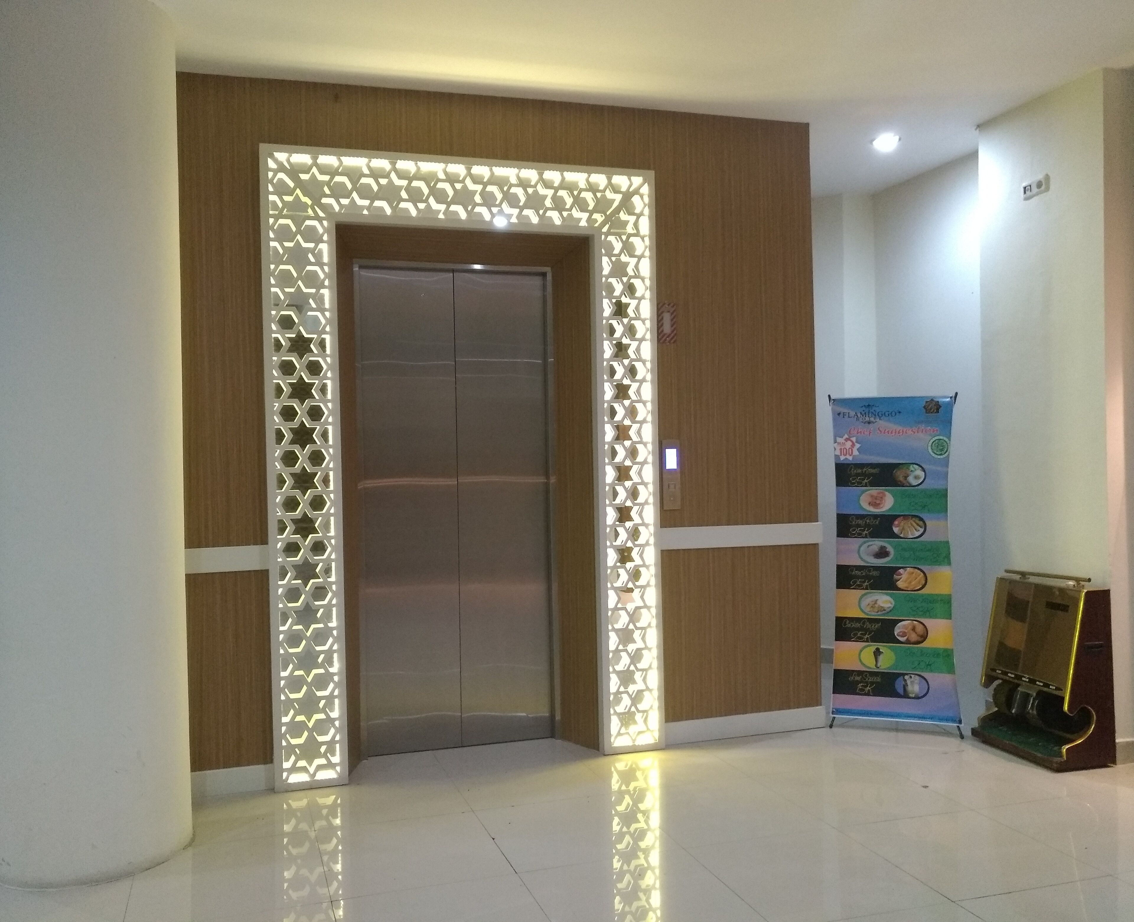 Un ascensor moderno con una lámpara geométrica encima, un panel de pared de madera y un tablero de menú digital a la derecha.