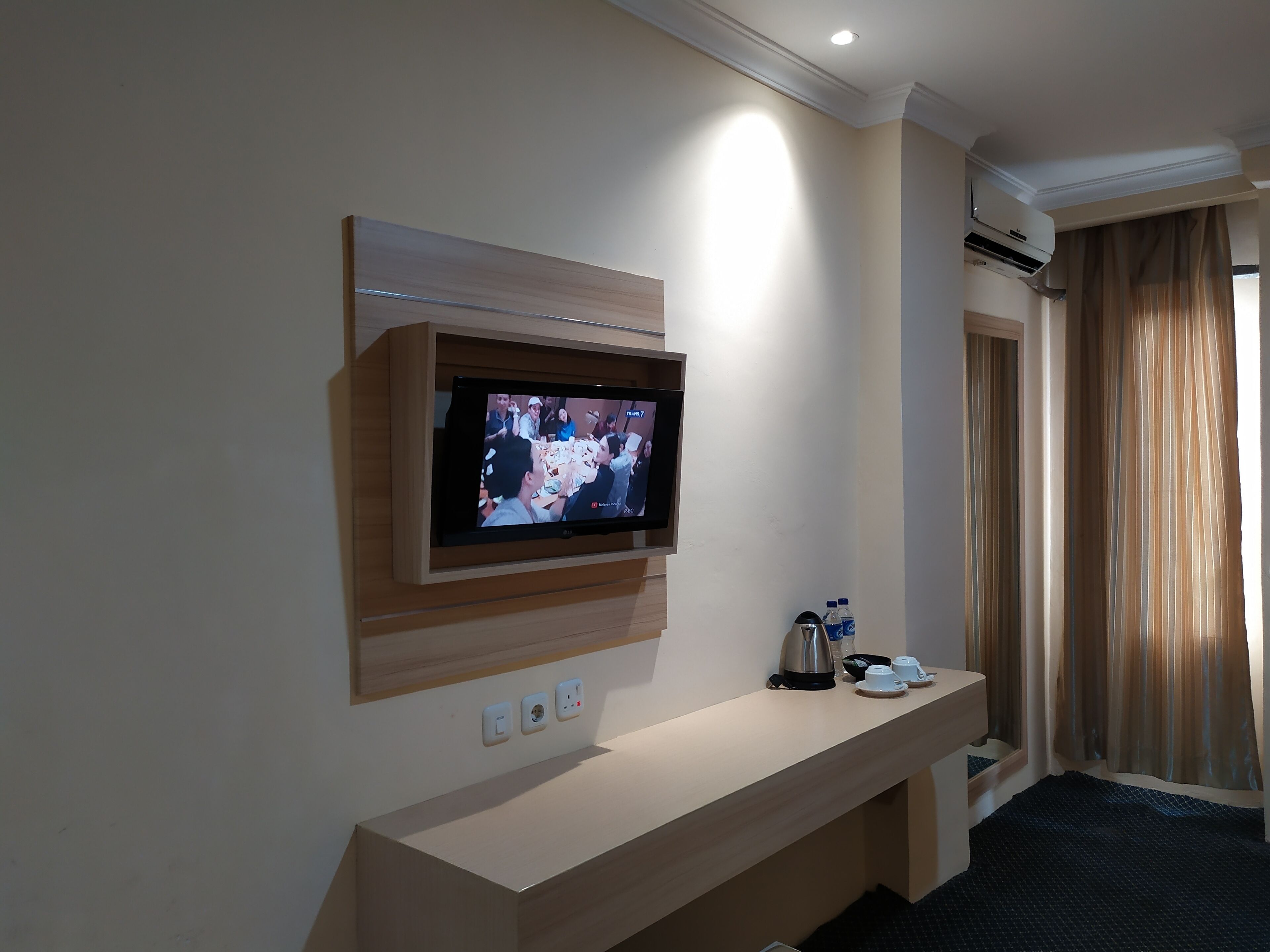A hotel room with a mounted TV, a bedside table with a kettle, cups, and a bottle, and a window with curtains.