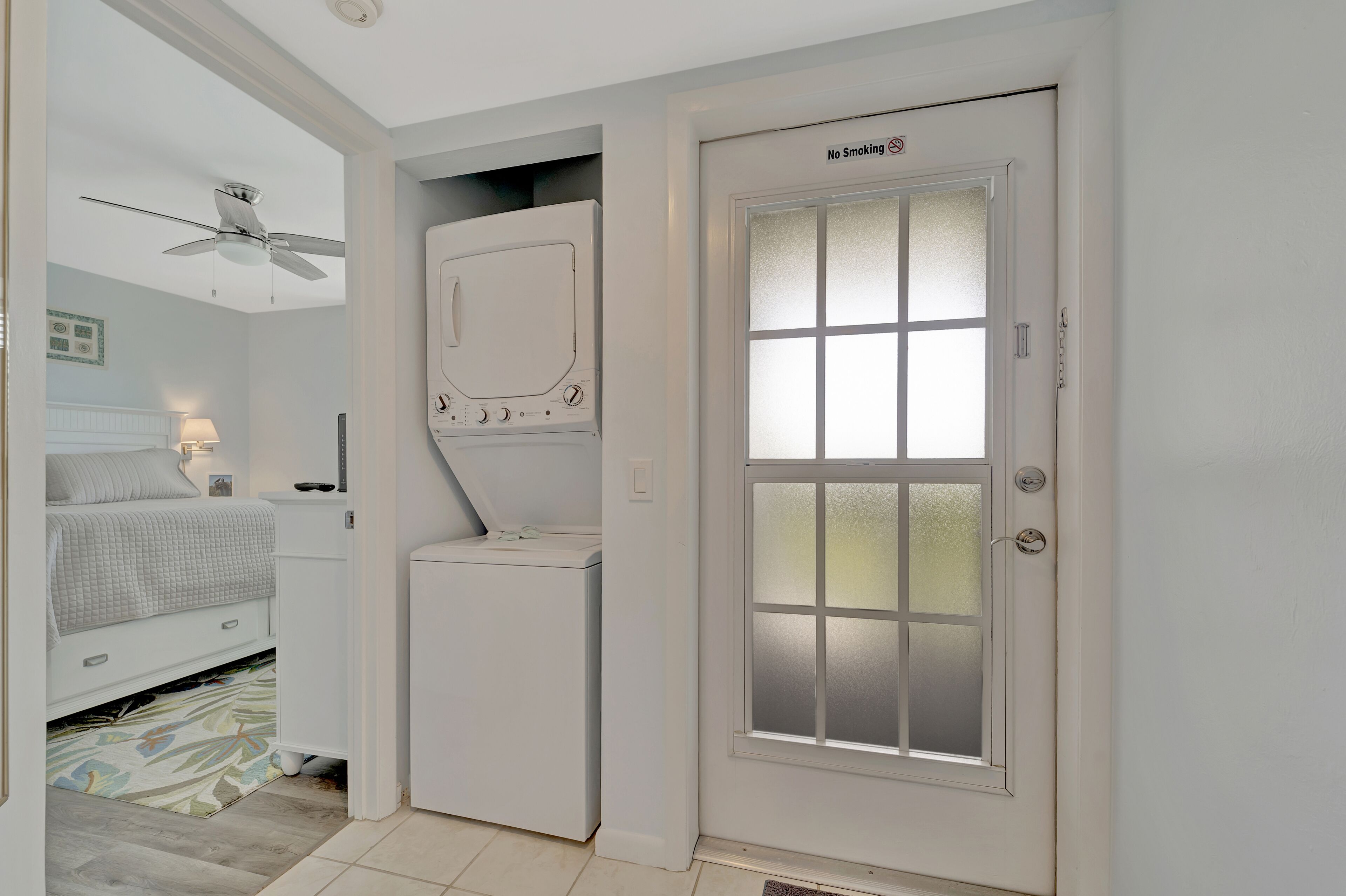 A compact bedroom with a bed, a ceiling fan, a washing machine, and a frosted glass door with a "No Smoking" sign.