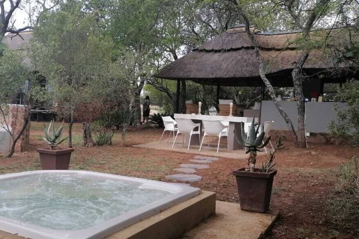 Un jacuzzi en un jardín con una mesa y sillas, rodeado de plantas en macetas y una estructura con techo de paja.