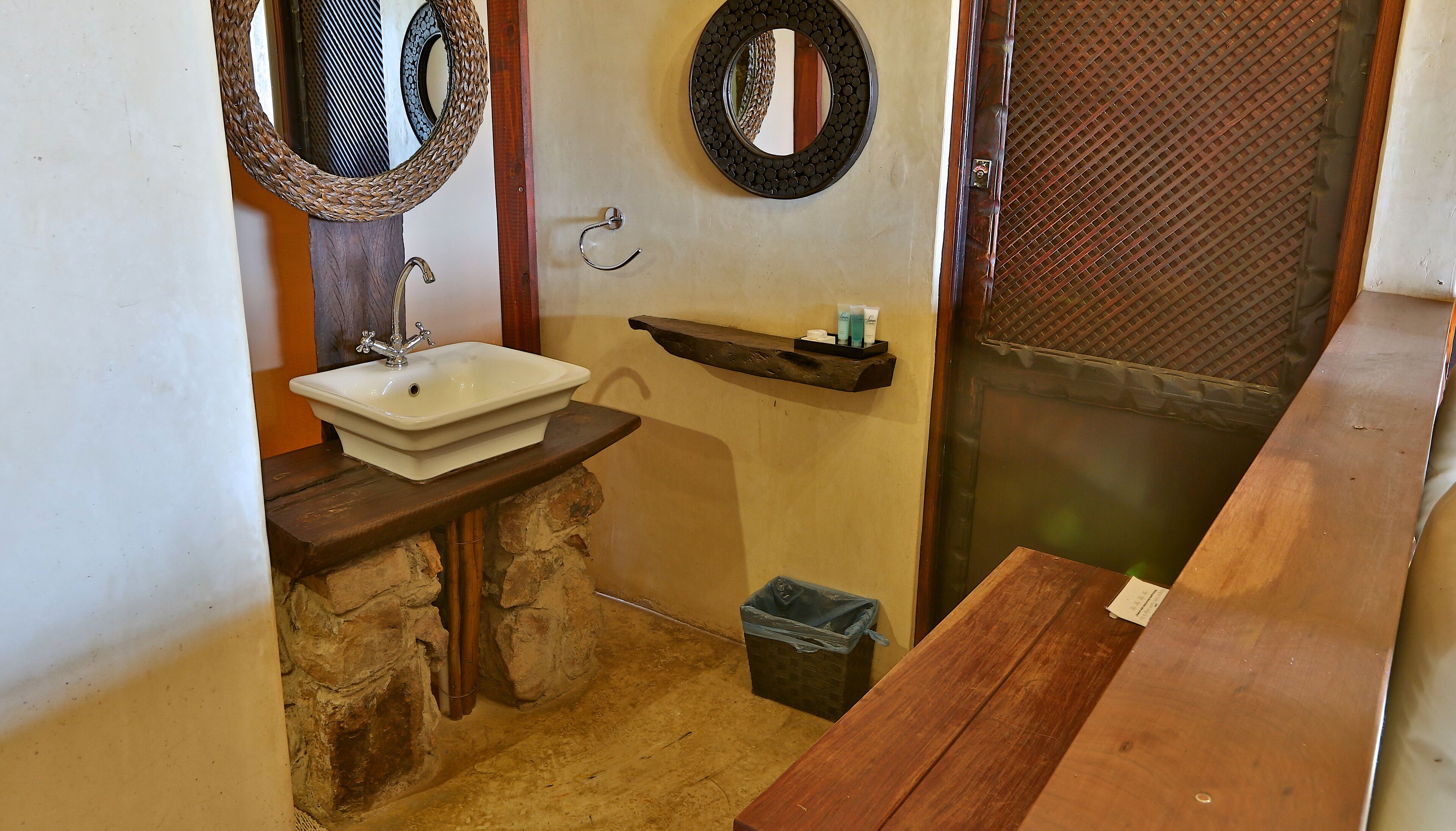 Une salle de bain avec un lavabo en pierre, deux miroirs fixés au mur, une étagère en bois contenant des articles de toilette et une poubelle.