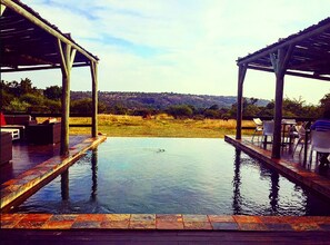 An infinity pool with a view of a grassy field and hills. There are two covered seating areas with chairs and tables. - Mangwa Valley Game Lodge & Spa (Hammanskraal)