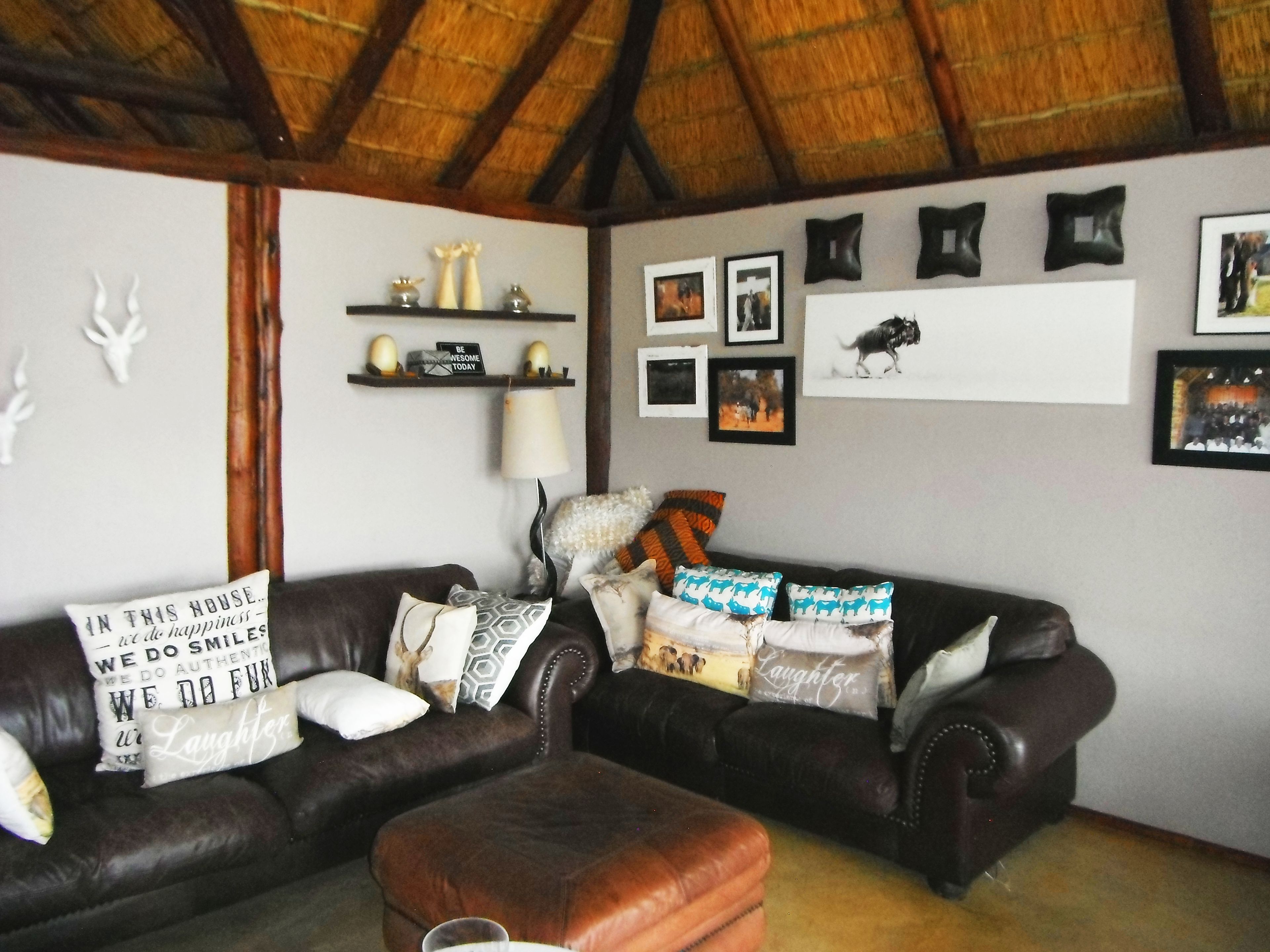 Una sala de estar con dos sofás de cuero oscuro, un reposapiés marrón y un techo de paja. Se observan estantes montados en la pared con objetos decorativos e imágenes enmarcadas.