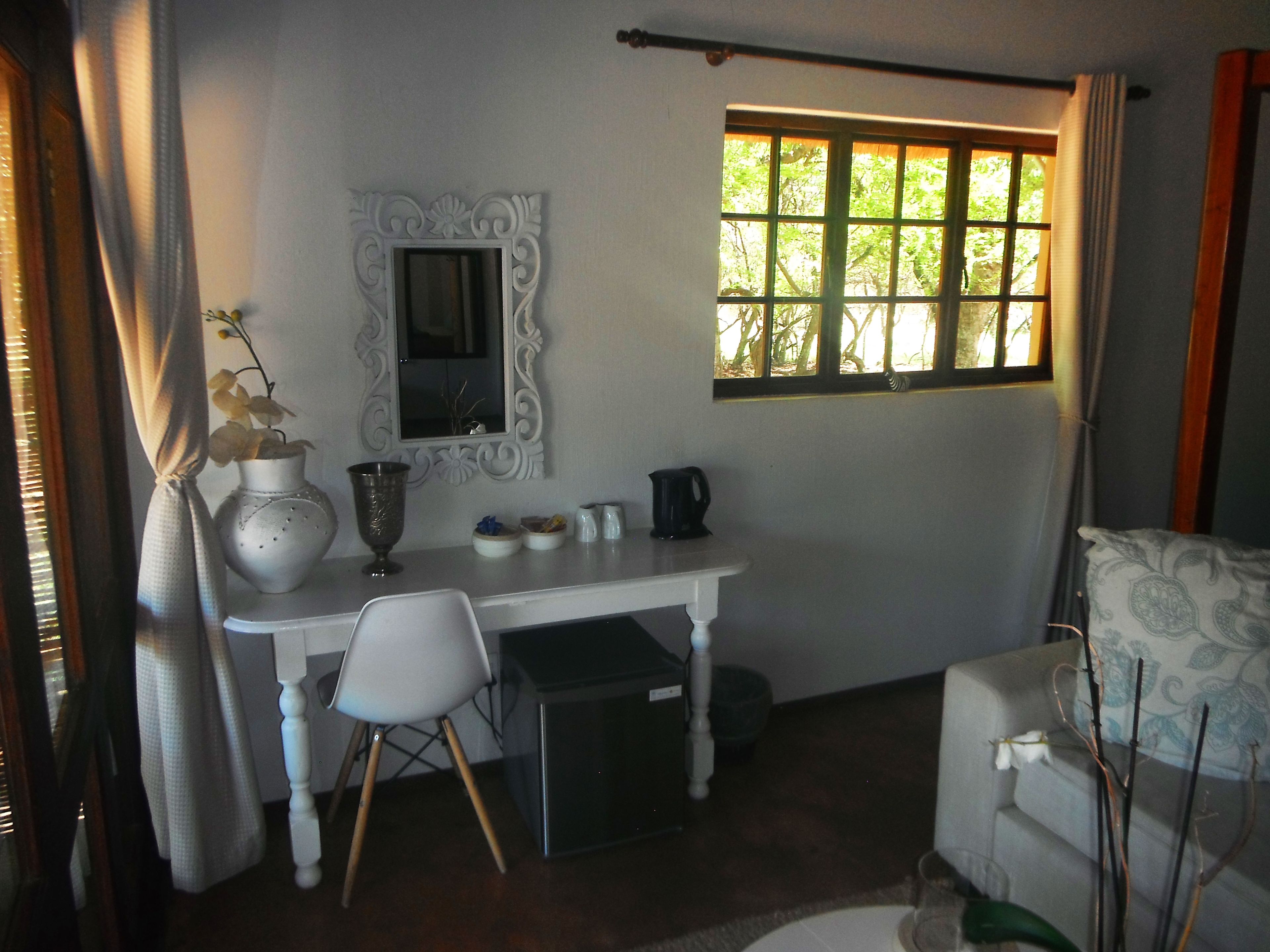 Una habitación pequeña y ordenada, con un escritorio blanco, una silla, un ventanal con vistas a la vegetación y una pared blanca con un espejo decorativo.