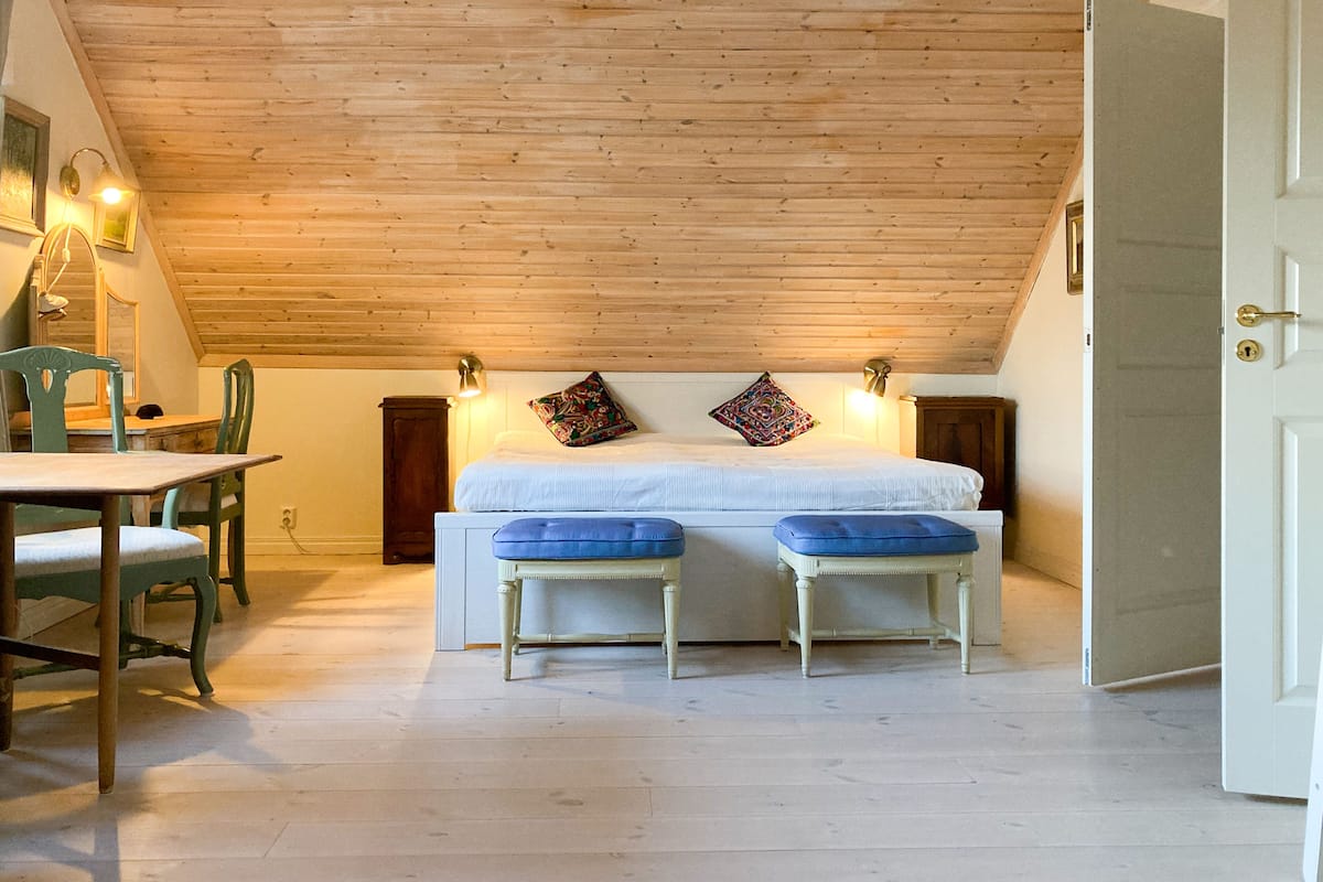 Un dormitorio acogedor en el ático con techo de madera, una cama con cojines azules y una zona de comedor con mesa y sillas.