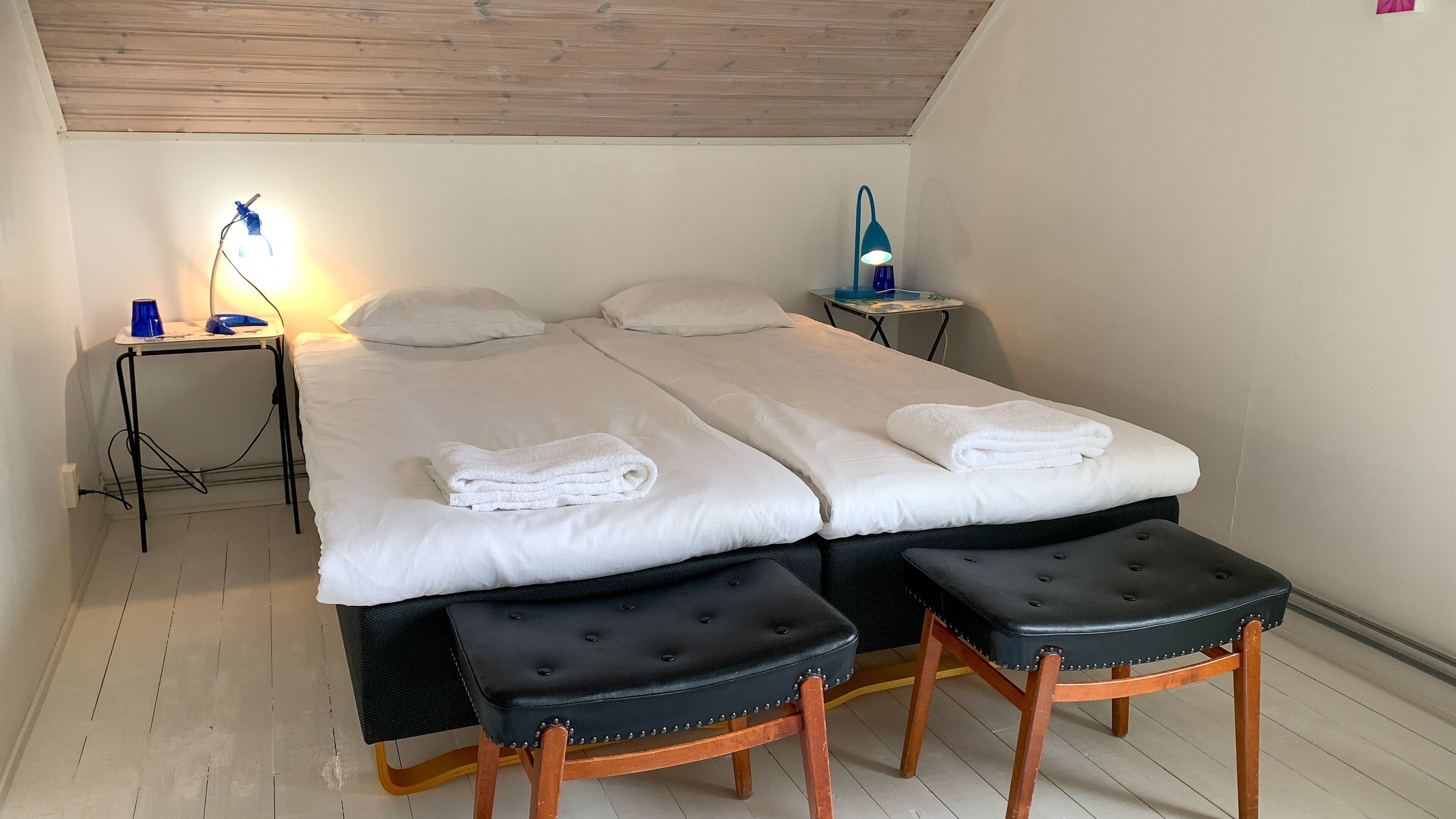 A neatly made bed with white linens and towels, a small bedside table with a lamp, a chair, and a wooden floor.