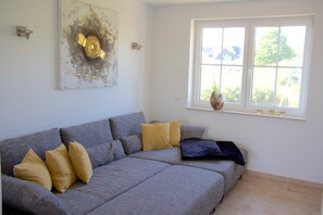 A grey sofa with cushions, a wall-mounted artwork, a window with a plant, and a thermostat.
