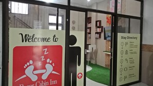 Welkomstbord voor Bogor Cabin Inn met een logo van een voetafdruk, een informatiebord met incheckinstructies en voorzieningen, en een blik op een kamer met een groen tapijt en een televisie.