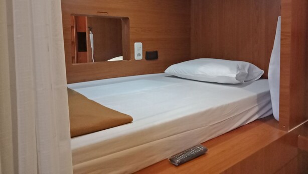 A single bed with a white mattress and pillow, a wooden headboard, a bedside table, and a remote control on the floor. - Bogor Cabin Inn - Hostel (Bogor)