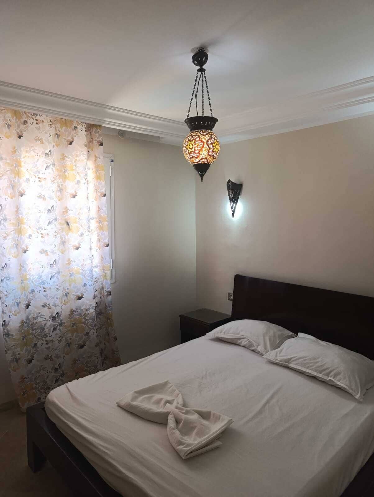 A bedroom with a bed, a nightstand, a wall-mounted light, and a window with a floral curtain.