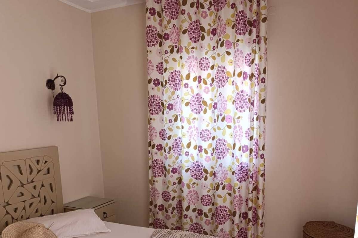 A bedroom with a bed, a nightstand, a wall-mounted lamp, a woven basket, and a window with floral curtains.