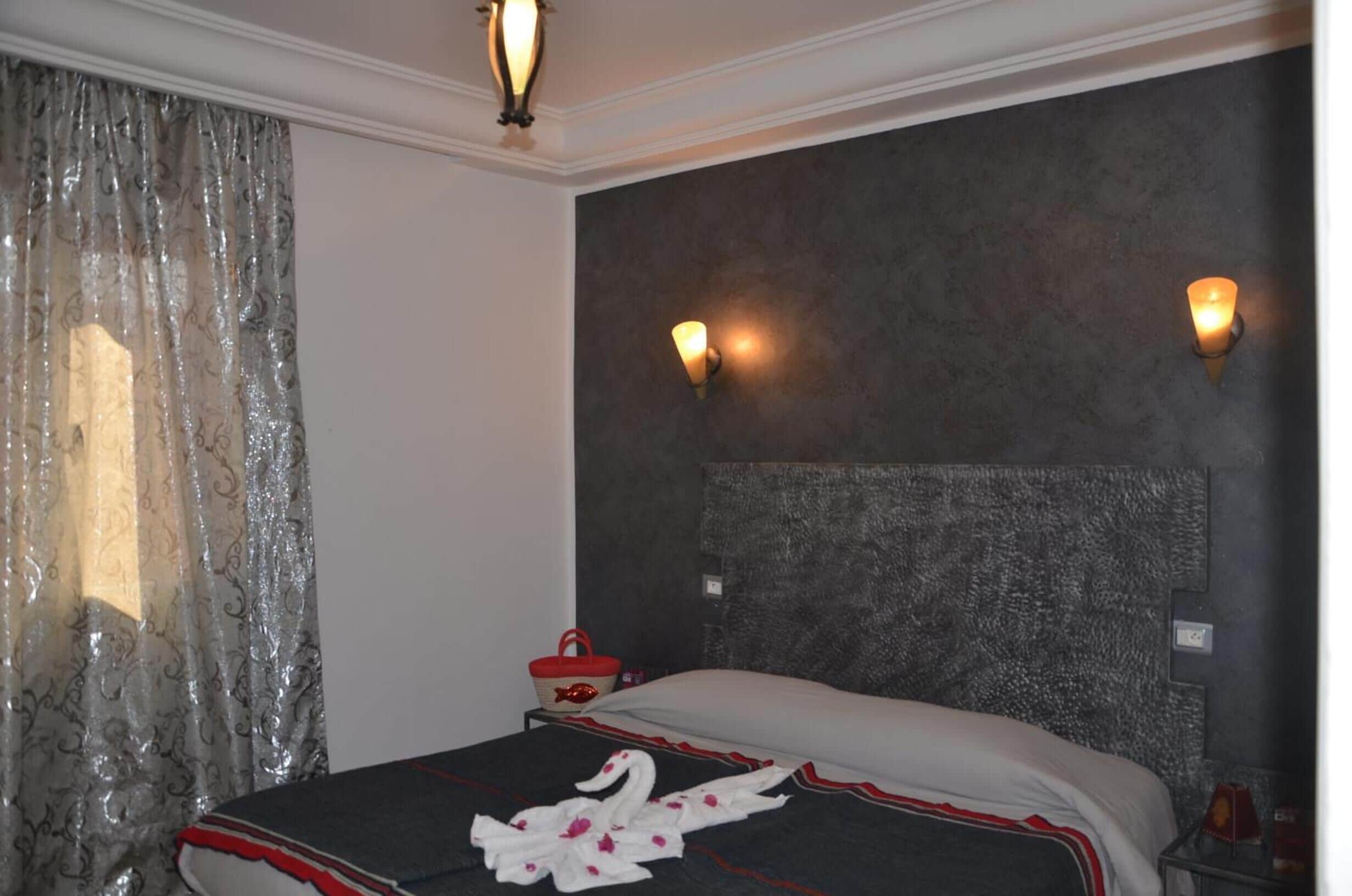 A hotel room with a bed, a headboard with wall-mounted lights, a red basket, and a window with sheer curtains.