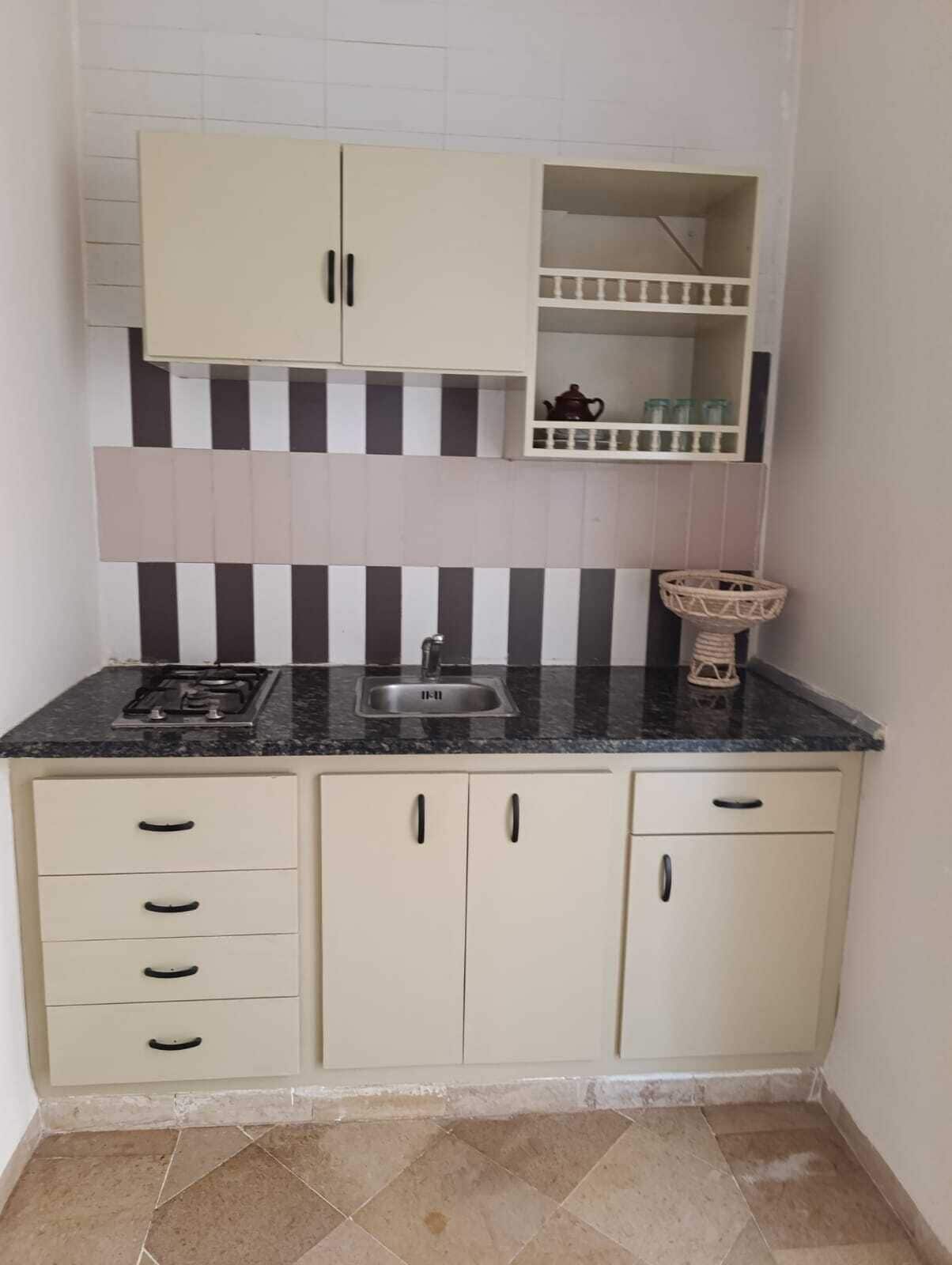 A compact kitchen with a gas stove, a sink, and a small shelf with a teapot and jars.