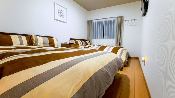 A hotel room with two beds, a bedside table, a window with curtains, a wall-mounted TV, and a light switch. - 73BNB House in Chiyozaki (Osaka)