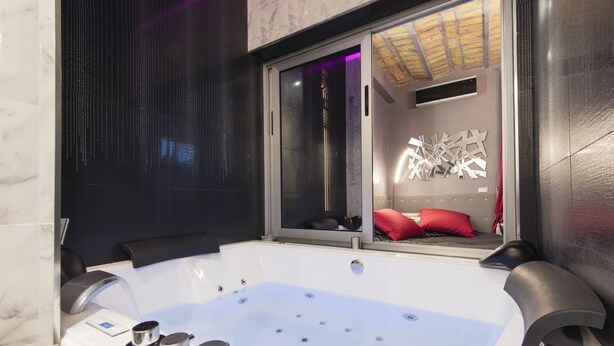 A modern bathroom with a large bathtub, a glass-enclosed shower, and a visible bed with red pillows. - Suite Sweet Rome (Rome)