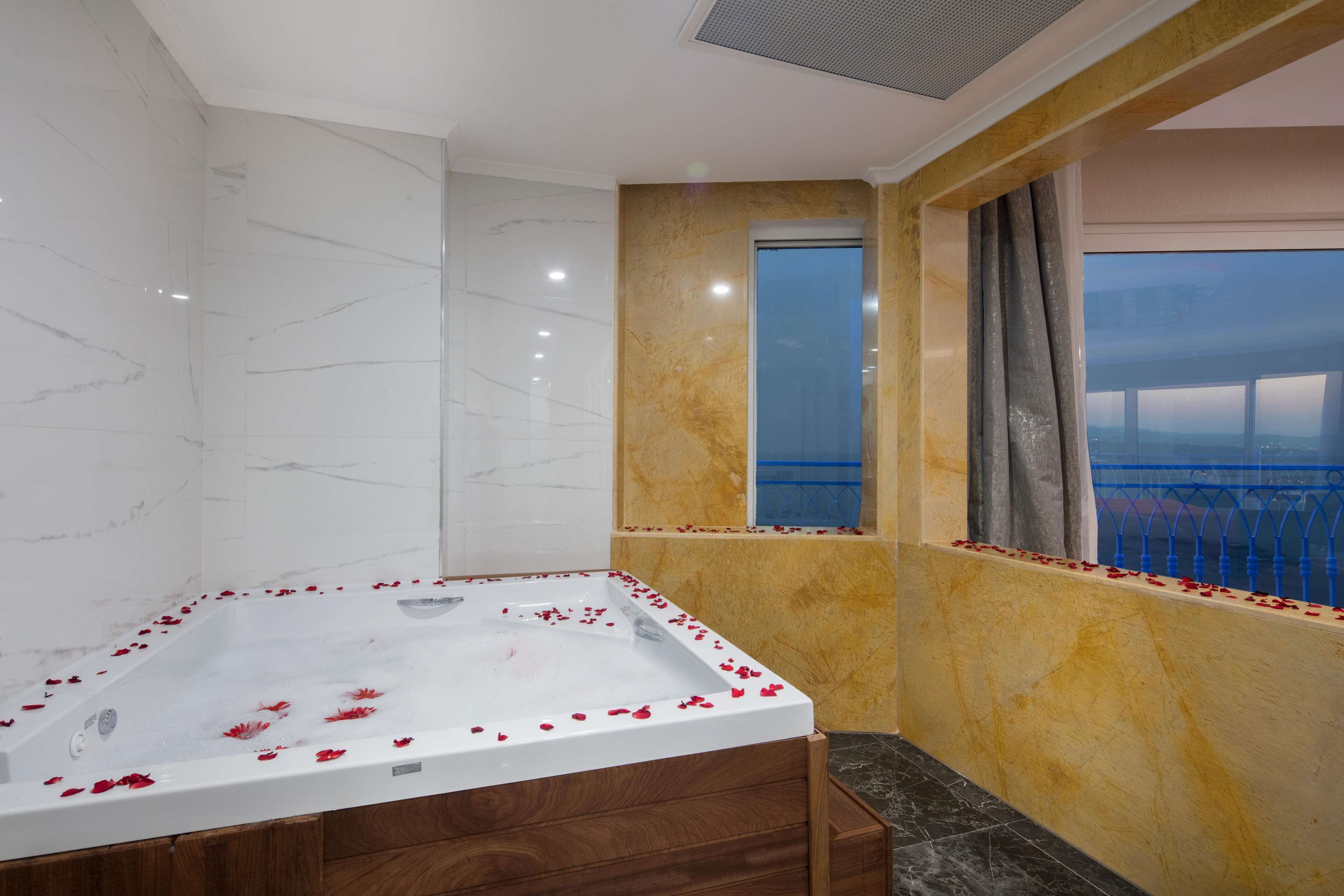 A bathroom with a marble wall, a bathtub with rose petals, and a view of the city through a window.