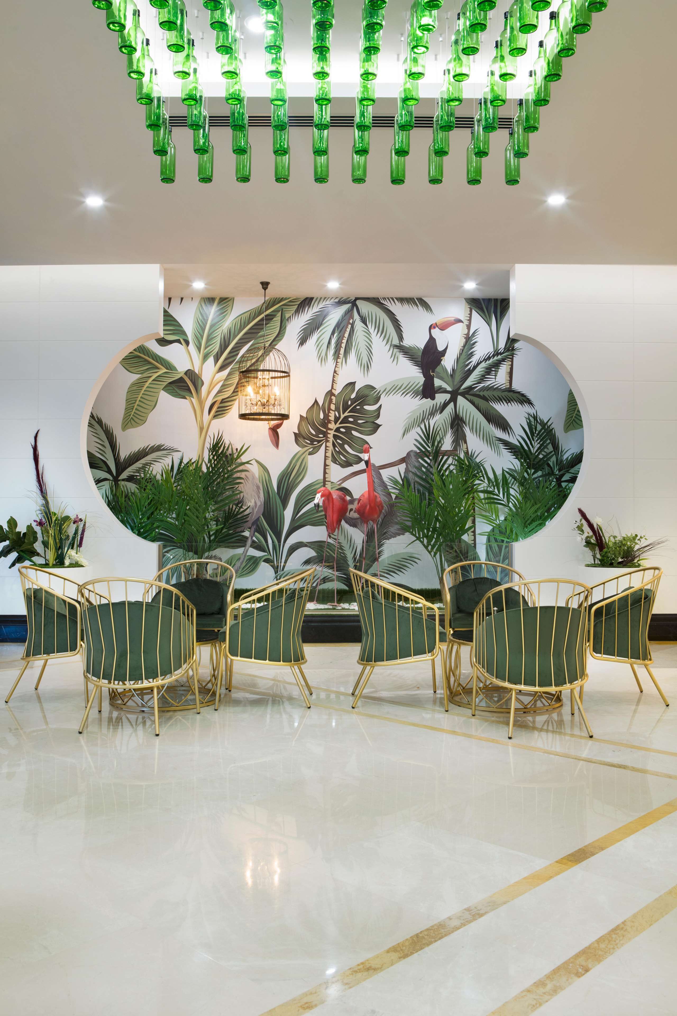 A lobby with a circular mural of tropical plants, green chairs, and a ceiling with hanging green bottles.