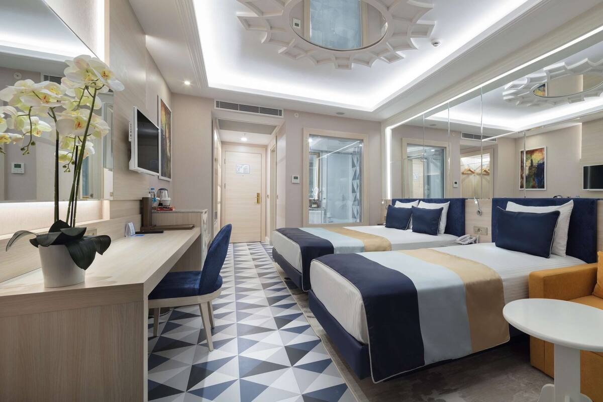 A modern hotel room with a desk, two beds, a sofa, and a small table.
