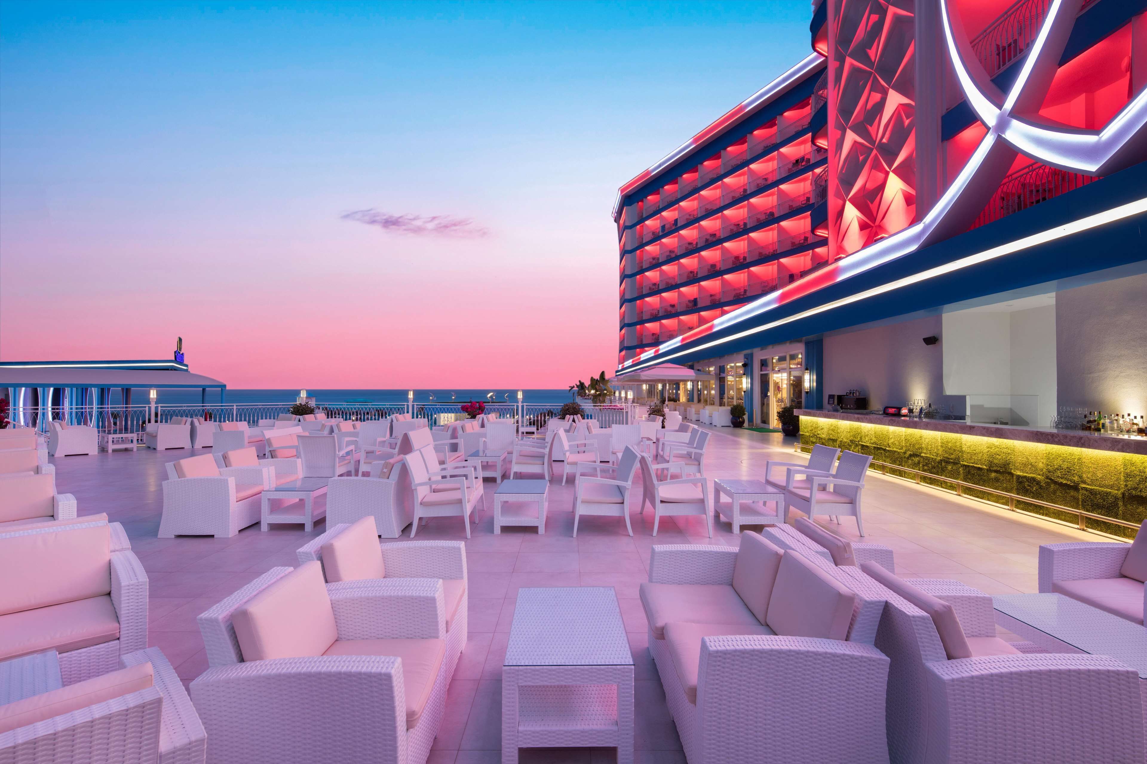 A rooftop bar with white wicker furniture and a large illuminated building in the background.
