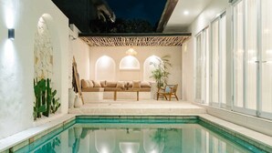 Un área de piscina interior moderna con una zona de estar, una pérgola de madera y un ventanal grande.