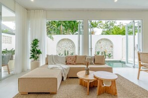 A modern living room with a beige sofa, two wooden side tables, and a large window overlooking a pool area. - Villa Putih Berawa (Canggu)