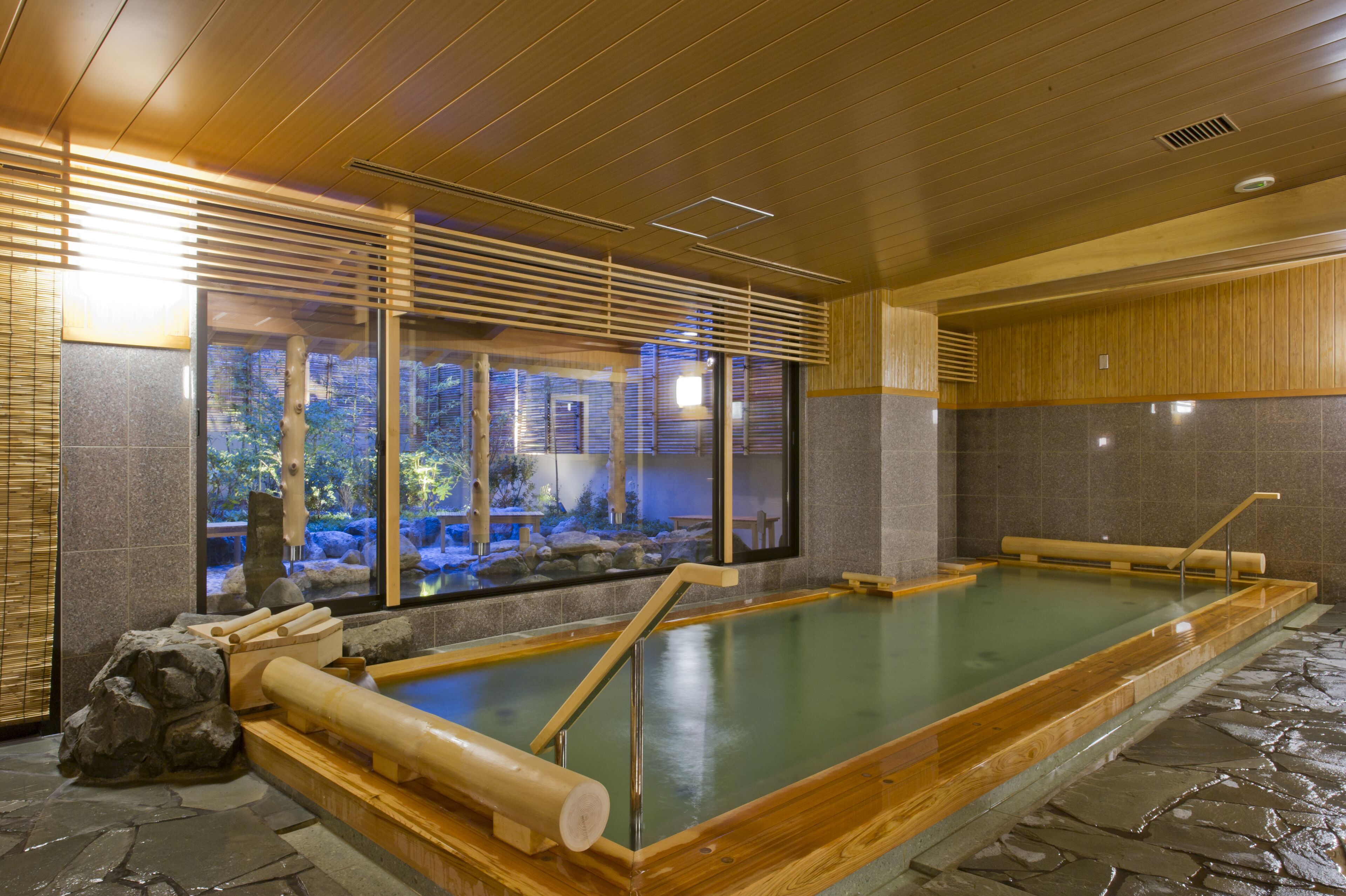 A traditional Japanese onsen with wooden benches, a stone floor, and a view of a natural landscape through large windows.