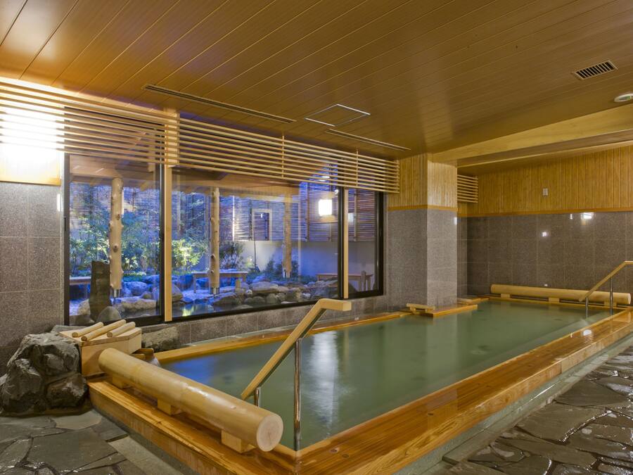 A traditional Japanese onsen with wooden benches, a stone floor, and a view of a natural landscape through large windows.