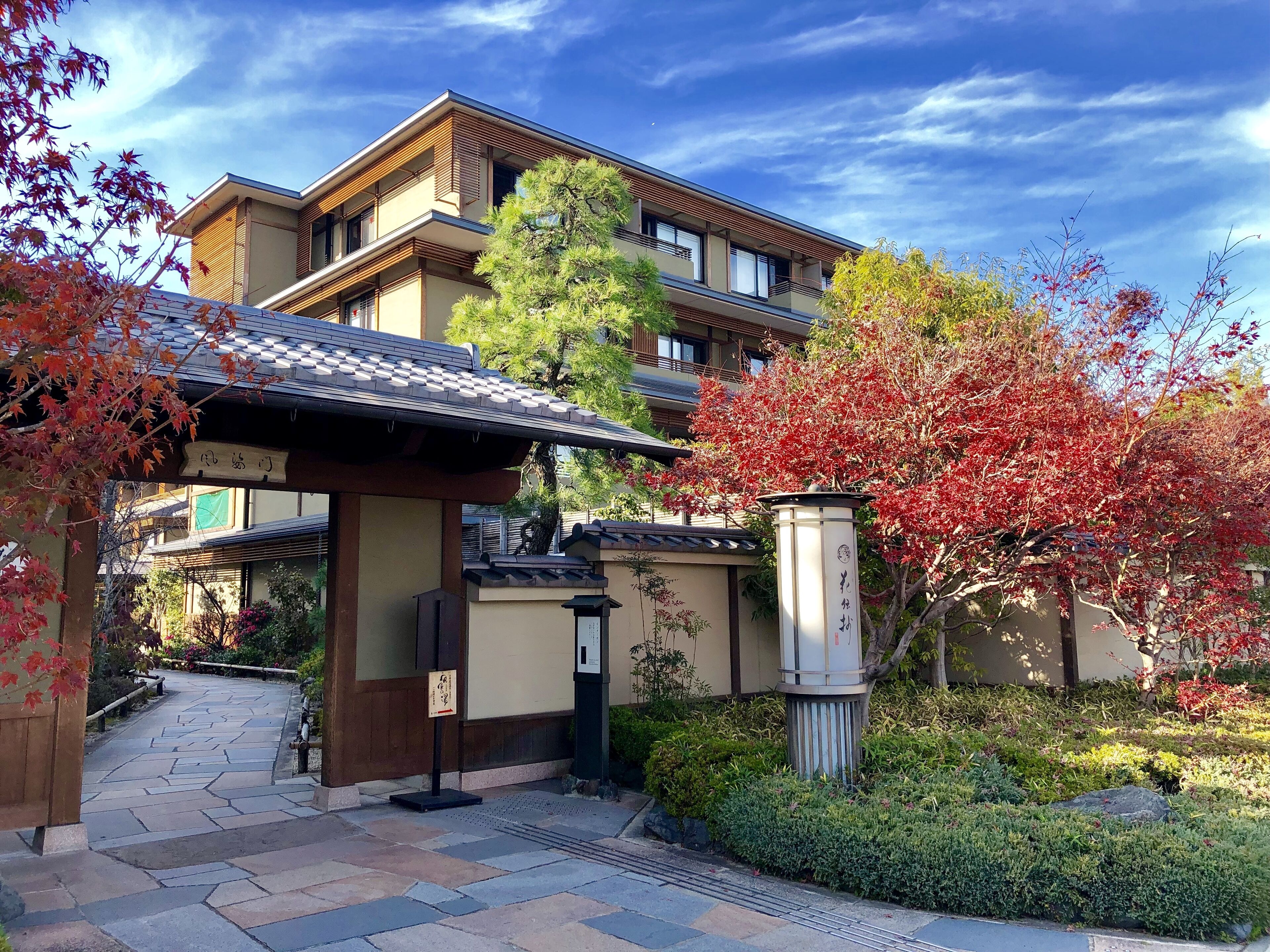 A traditional Japanese building with a tiled roof, a stone pathway leading to the entrance, and a well-maintained garden with red foliage.