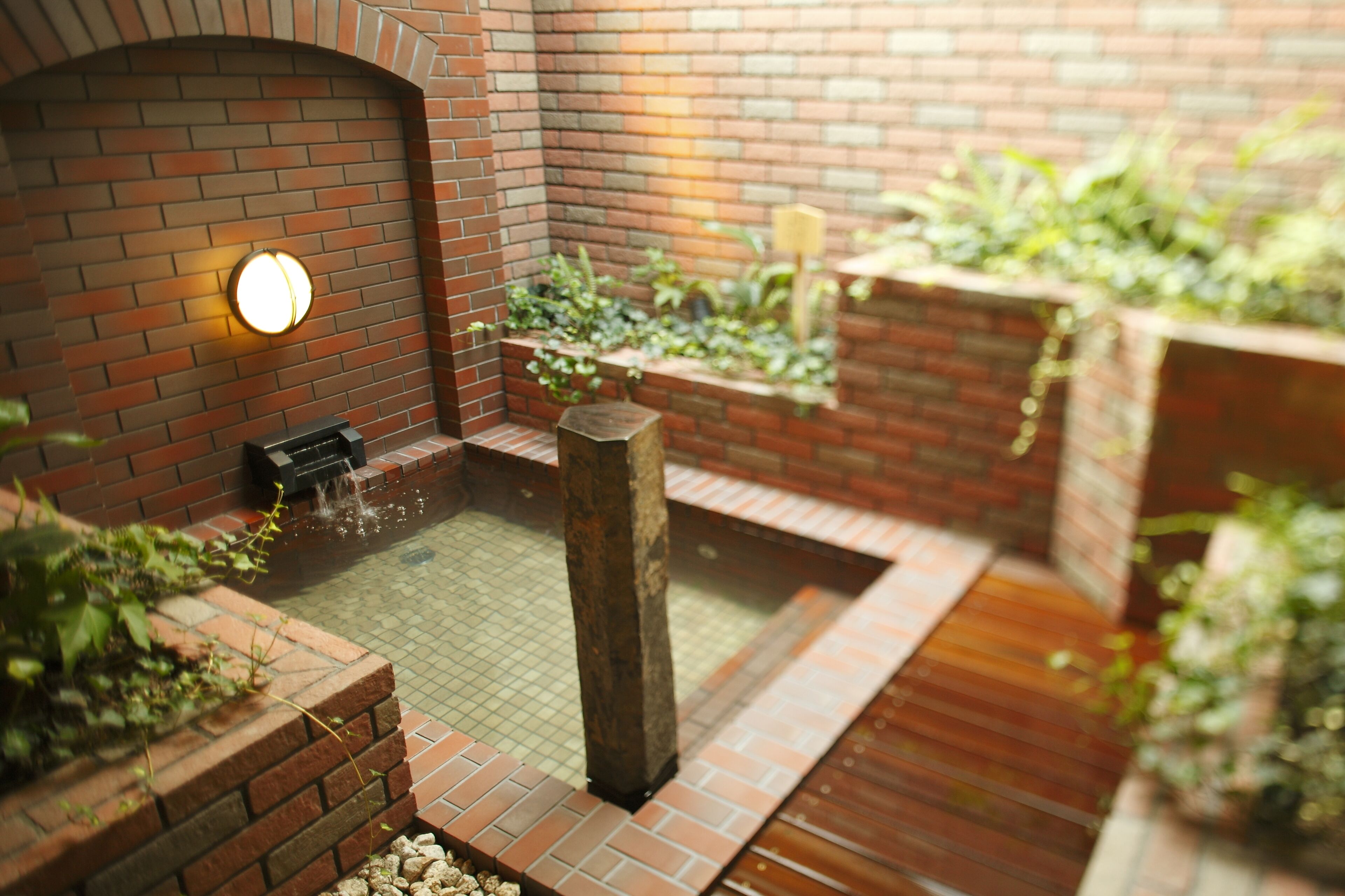 A brick-walled courtyard with a small water feature, a wooden bench, and potted plants.