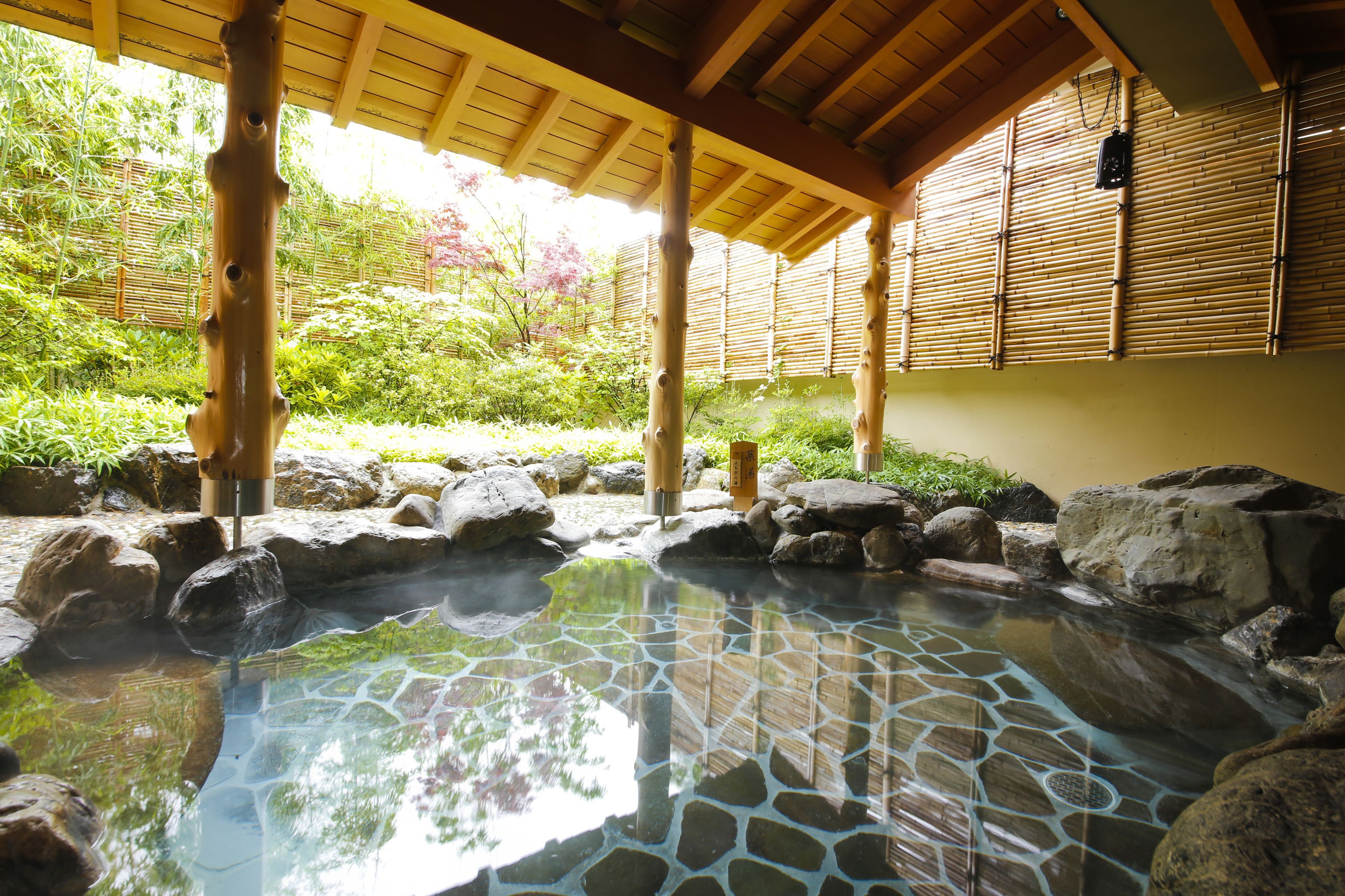 A traditional Japanese onsen with a stone-tiled hot spring, wooden pillars, and a bamboo backdrop.