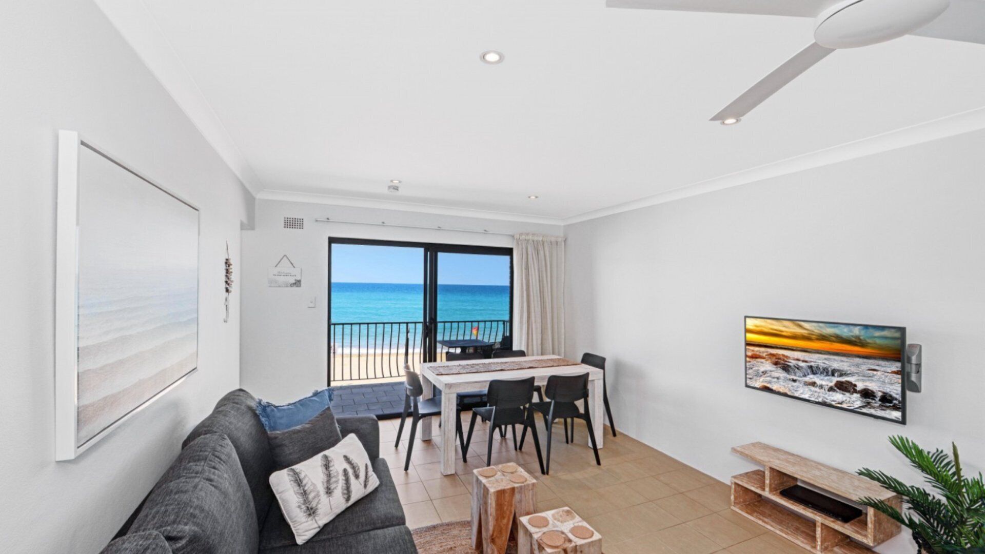 A modern living room with a balcony overlooking the sea, a dining area, and a mounted TV.