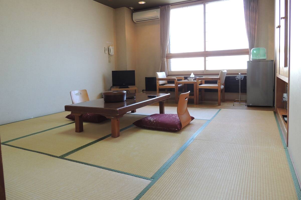 Een traditionele Japanse kamer met tatami vloerbedekking, een lage tafel met kussens, een televisietoestel en een koelkast.