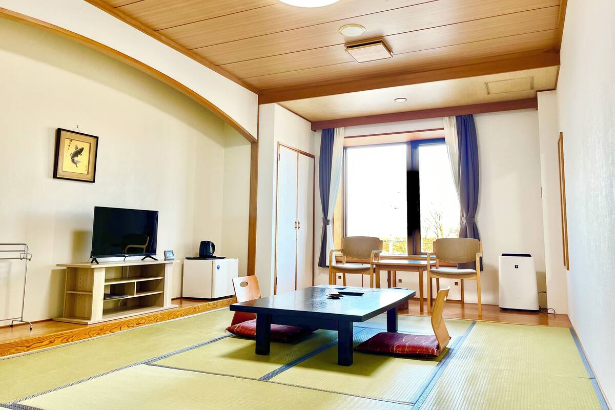 Een kamer met een tatami vloer, een televisie, een kleine koelkast, een tafel met kussens en een raam met gordijnen.